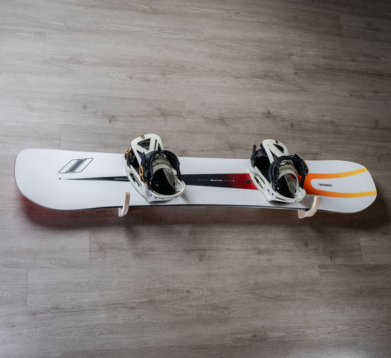 Single wooden snowboard wall rack being holding a white snowboard with white bindings, on a grey wooden wall. 