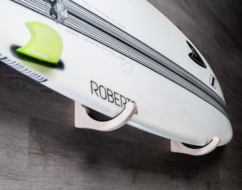 Close up view from the side of the single wood wall rack for surfboards, holding a white shortboard.  The rack is mounted to a grey wooden wall. 