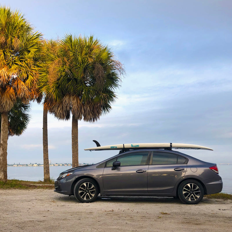 Dark grey honda 4-door sedan shown parked next to a waterway while a white Stand Up Paddleboard is mounted to the roof using Epic Suction-Mount Roof racks. There are a few palm trees next to where the car is parked. 