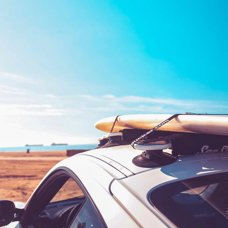 Universal suction-mount roof rack shown on a white sports car parked at the beach.  There is a surfboard strapped to the board rack on the roof.  A long strand of sand is in the distance with the ocean and some ships out to sea
