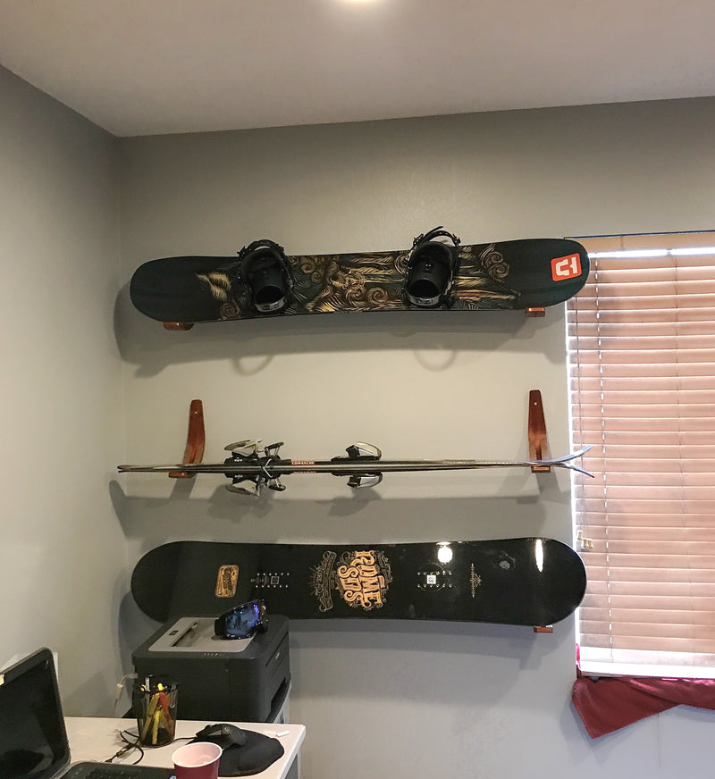 Three Surfboard, Ski, Wakeboard, snowboard Wall rack shown holding two snowboards and one pair of skis in a bedroom. The wall is light grey and the racks are mounted next to a desk.