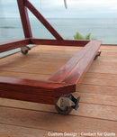 Detail shot of the custom tower rack in brown mahogany, showing the locking wheels attached. There is a wave breaking in the distance.