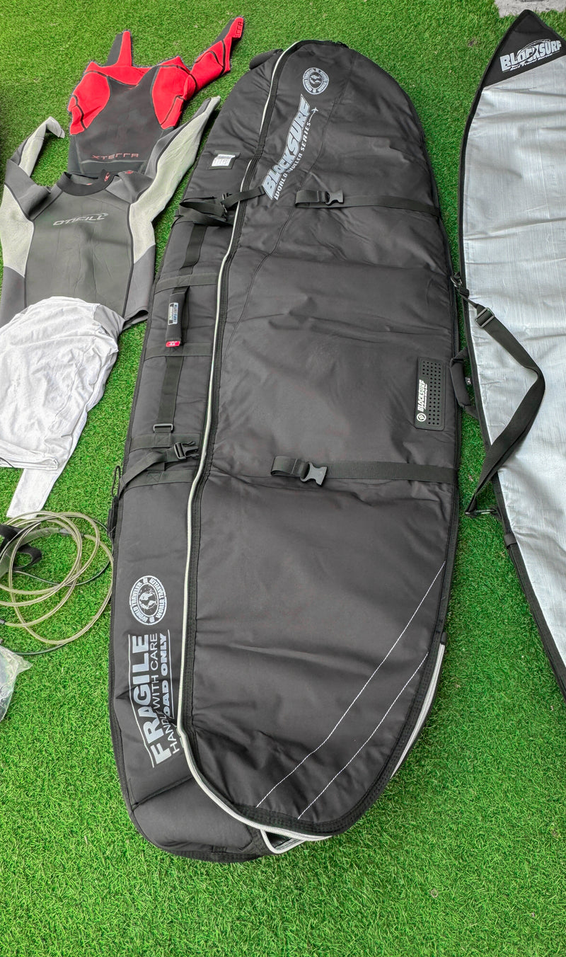 A Black surfboard travel bag is laid out on some fake grass with wetsuits laid out next to it.