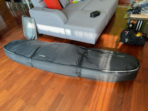 Surfboard travel board bag shown in a living room on wooden floors. The board bag is packed with multiple surfboards awaiting airline travel. A grey couch is in the background.