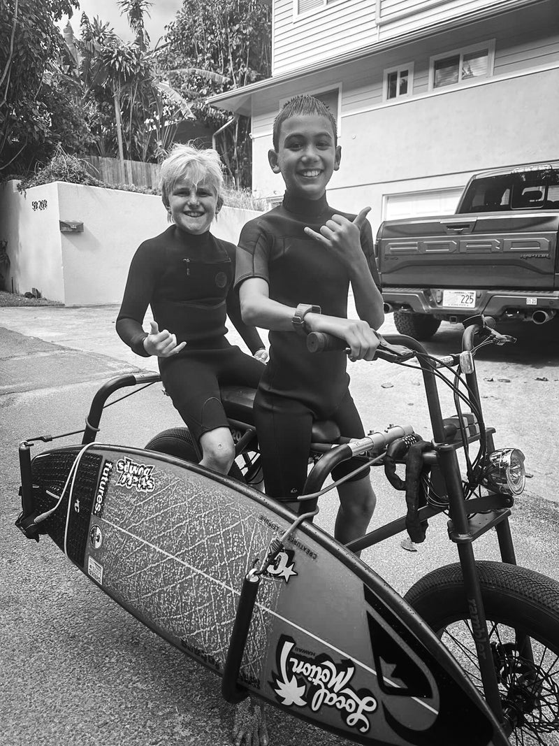 Two kids on an e-bike electric motor driven bicycle with a surfboard attached.  The kids are in wetsuits are smiling and giving the shaka gesture with their hands.