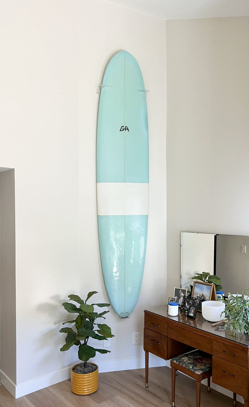 Clear Acrylic vertical surfboard wall rack shown holding a blue fun-shape board in a bedroom in front of a dresser.