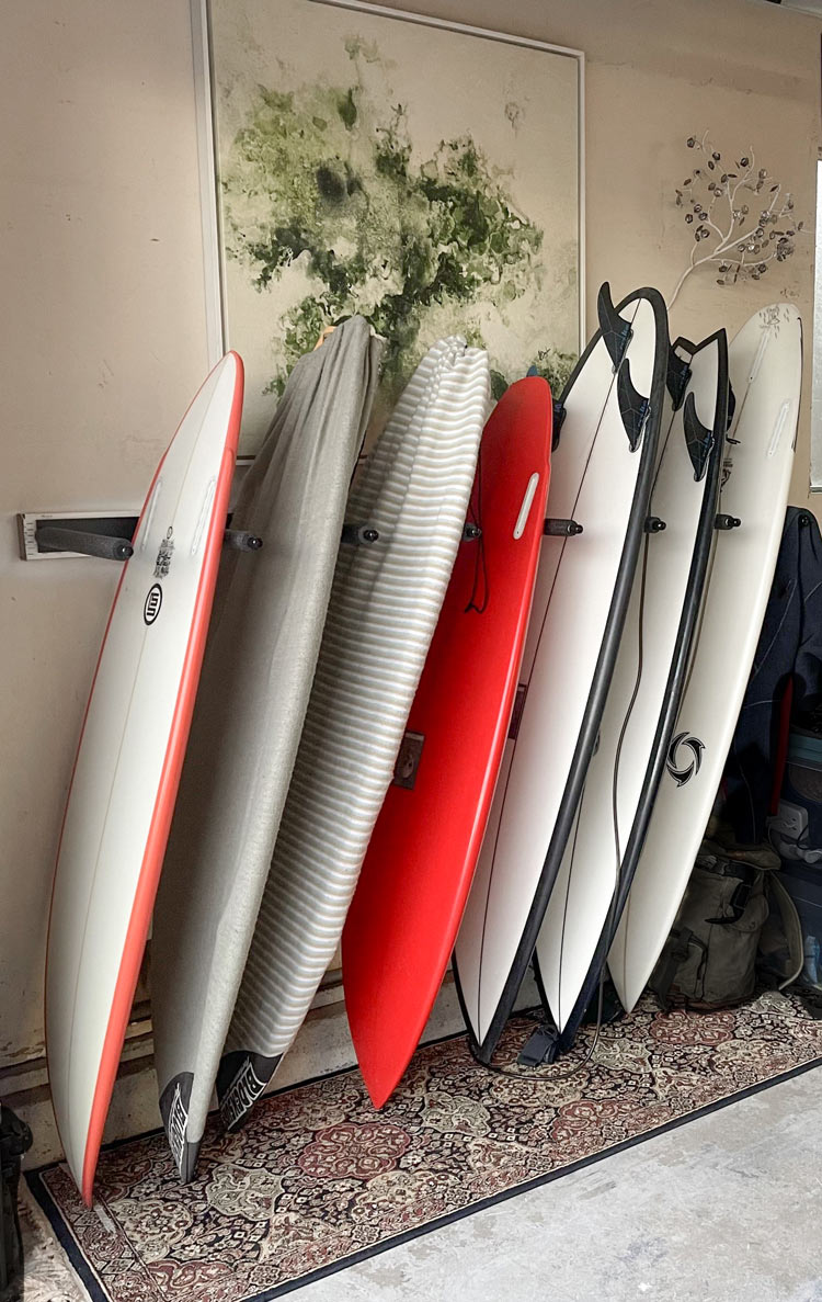 Vertical surfboard wall racks in black metal shown holding several high performance shortboards (surfboards) in a living room. There is a Persian rug below the surfboards protecting the bottoms of the boards.