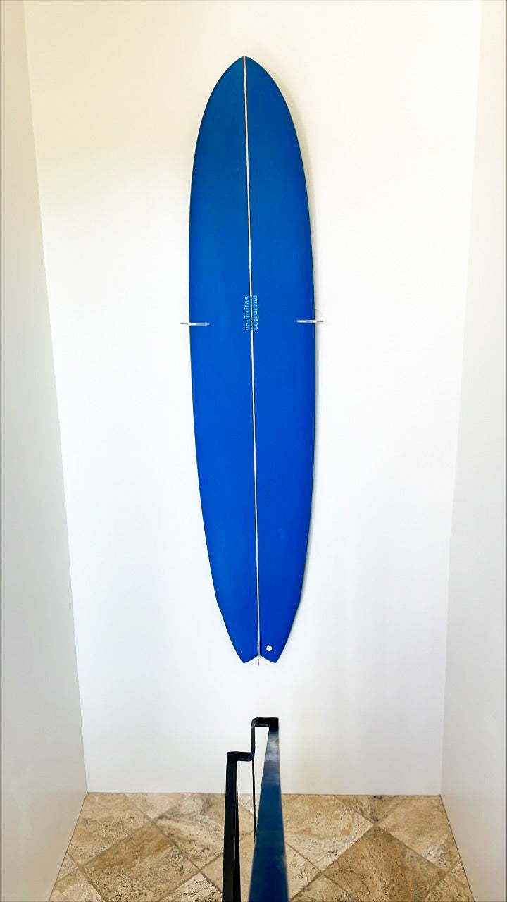 Blue surfboard shown mounted in a stairwell using the clear acrylic vertical wall rack. There is white walls, and marble floors, with a black steel handrail in the foreground.