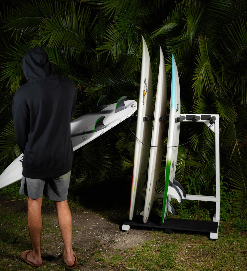 Marshmellow foam freestanding surfboard rack holding multiple surfboards in front of some palm plants, while a surfer holds a board and walks off away from it. 