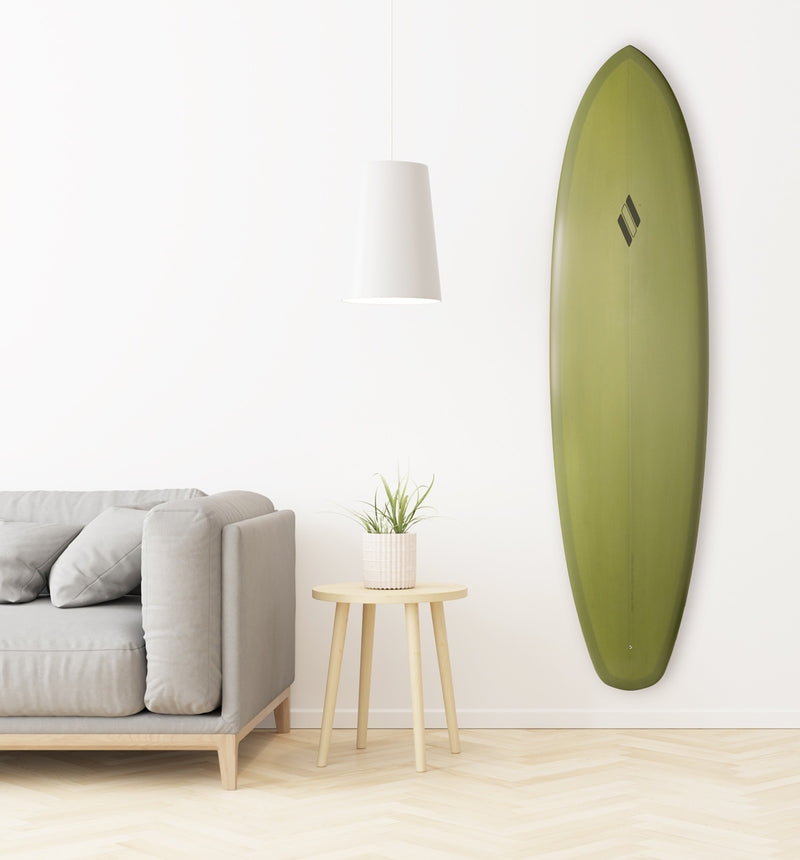 Acrylic vertical surfboard wall mount holding a green surfboard, shown mounted in a living room next to a grey couch and wooden end table with a plant.  The floor is wood and the wall in the background is white.  