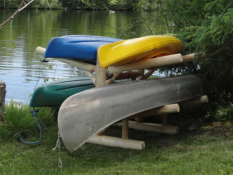Lakeside Kayak Rack holding 3 Kayaks and one Canoe right by the water.  Tucked up in a bush sitting on beautiful green grass with water in the background, glistening ever so slightly. 