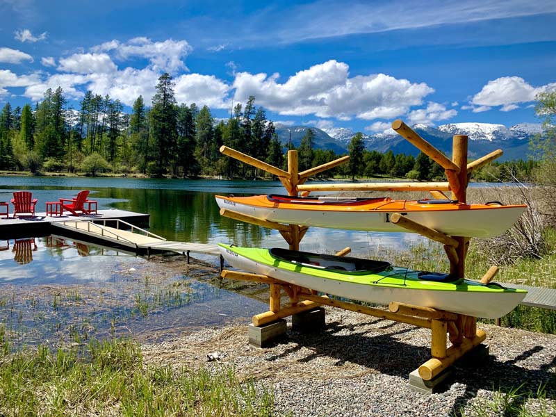 6 Log Kayak Rack holding 2 kayaks one orange and one green.in front of a dock on a beautiful lake with clouds and mountains in the background.  Red Chairs in the foreground.