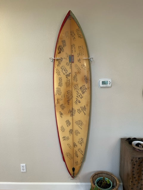 vertical shortboard acrylic wall rack shown holding a customers surfboard that has a bunch of signatures on it. 