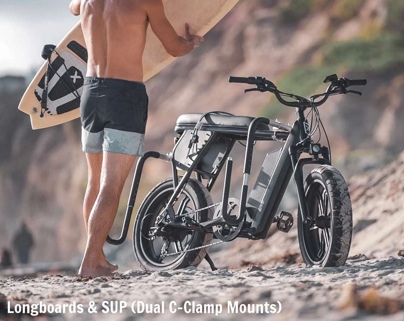 Longboard e-bike rack being used at the beach next to a cliff, with a surfer coming out of the ocean with his surfboard.  White text says Longboards & SUP (Dual C-Clamp Mounts)