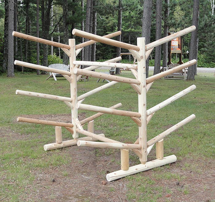 8 Slot watercraft freestanding storage rack made of wooden logs with no boats in it.  Sitting on grass in the woods with some chairs in the background. 