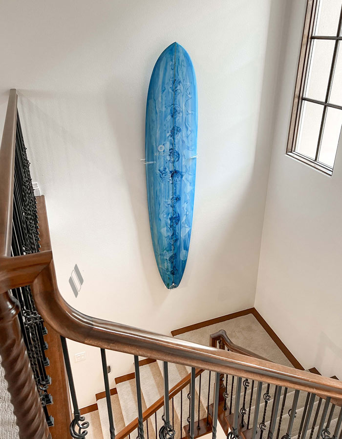 Shortboard version of the clear acrylic vertical surfboard rack is being displayed going down the stair well. Beautiful woodwork is done on the banister, as a blue longboard is being held up in the middle of the wall going down the stairs.