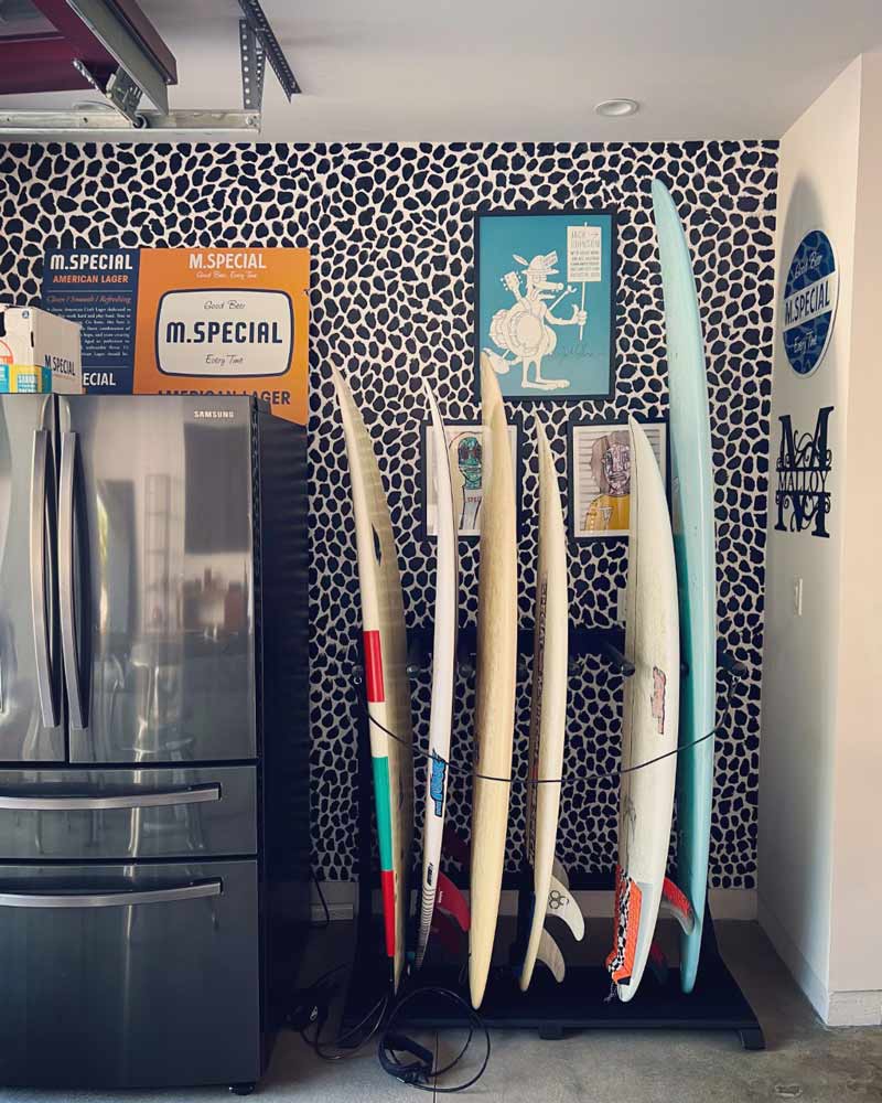 Blackout surfboard rack shown holding several shortboard surfboards and a longboard.  Has a black and white cheetah print on the wall in the background, and a stainless steel fridge to the side. 