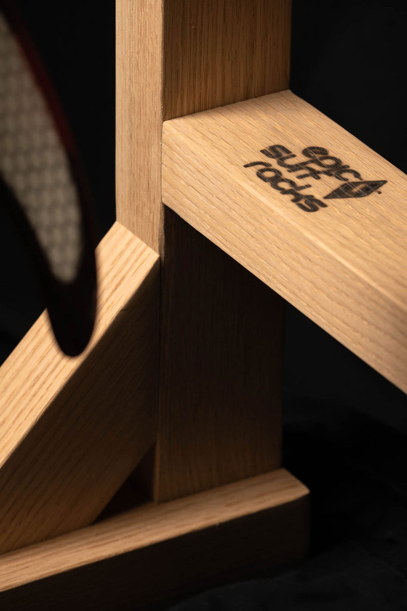 Close up image of the bottom of the surf rack. The Epic Surf Racks logo is burned in red-oak hardwood rack.  There is a fin in the foreground of the image. 