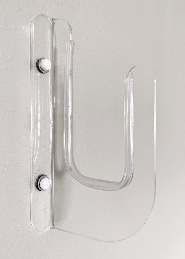 Close up image of the acrylic flush mount surfboard rack.  The surf rack is mounted to a grey wall, using two bolts, that have white caps.   The rack is entirely clear. 