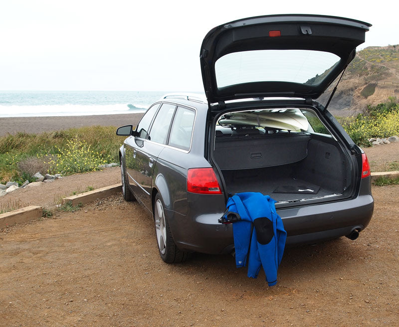 SeatRack car head rest surfboard rack shown being used in a station wagon parked next to the beach.  The car's hatch is open where you can see a wetsuit, and multiple surfboards being held on the rack. 