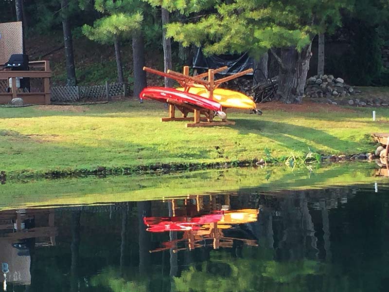 Shore side Kayak rack displaying 2 kayaks on a green grassy knoll.  Water has a mirror like texture with green trees in the background.