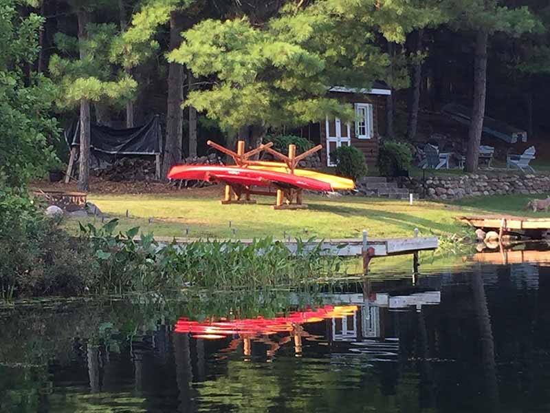 Lake House with a ray of light shining on a freestanding kayak rack with dark brown wood.  The rack is holding a red and also a yellow kayak.  Water in the foreground has a mirror-like finish showing some vegetation, and a dock leading from the grass to the water.  A small house is in the background tucked up behind some trees.  A beautiful sight. 