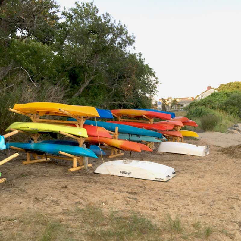 Showing multiple Kayaks being stored at a beach club using several of the 6 SUP and Kayak Log Storage Rack.  Buried in the Sand.  Safely out of harms way.  Green Trees and some houses in the background.