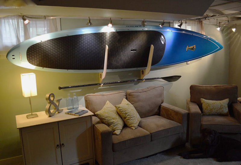 Single SUP Wall Rack shown in a living room holding a paddle over some couches and beautiful lighting.  A dog is shown in the corner looking at the camera