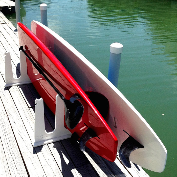 SUP & Surfboard Rack shown on a Dock holding 2 paddle boards and a paddle. One board is Red the other is white with green water in the background. The rack is white and is bolted to the pier.