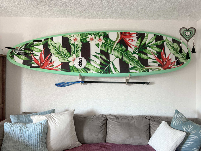 Stand Up Paddleboard Single board being displayed in a living room over a couch.  The board has a beautiful floral and striped design and a black and blue paddle is being held directly below on the rack. 