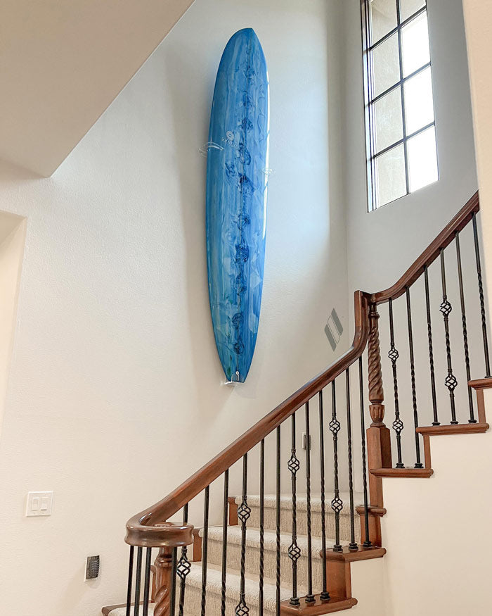 Shortboard version of the clear acrylic vertical surfboard rack being displayed in a beautiful stairwell. A blue board is displayed in the vertical position in the middle of the wall going up the stairwell. There is a window providing light to the right and the banister to the stairwell is a beautiful dark wood.