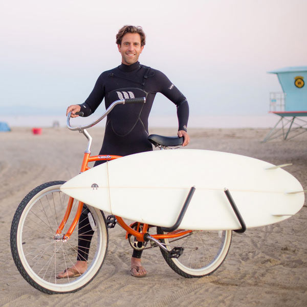 Single-Mount surfboard bike rack shown on the beach attached to an orange beach cruiser. A man in a wetsuit is behind the bicycle and is smiling.