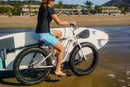 SUP Bike Rack shown mounted to a white beach cruiser while a girl rides the bike up the beach. There is a building and some palm trees in the background.
