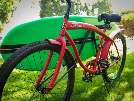 Single-Mount surfboard bike rack shown mounted to a red beach cruiser.  The bike rack is holding a green surfboard with stripes.  The bike is sitting on a lawn. 