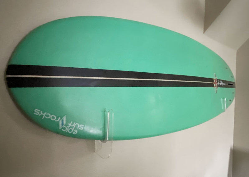 Acrylic flush mount surfboard rack shown holding a green surfboard with a black stripe.  The board is mounted pretty close to the wall, and the board has a sticker that says epic surf racks in the lower left corner of the board. 