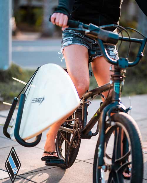 Front image of the single-mount surfboard bicycle rack. The rack is holding a single white Firewire surfboard.   A girl 's legs are shown while she sits on the bike. 