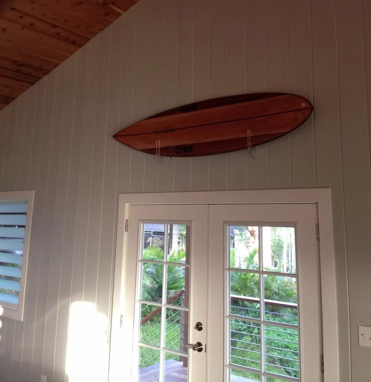 Wooden surfboard shown mounted using the acrylic flush mount surfboard holder above a doorway.  There is a wooden ceiling in the room, and a deck just beyond the glass doors. 